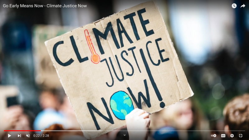 Screenshot of youtube video Go Early Means Now, a song Edna from Tāmaki Makaurau wrote to support XR's open letter to Prime Minister Ardern. Screenshot is showing a protest sign saying Climate Justice Now.